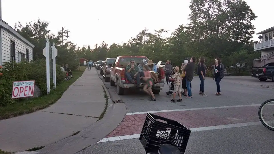 People gather beside a red pickup truck on a street at dusk near a sign advertising an open museum and gift shop.