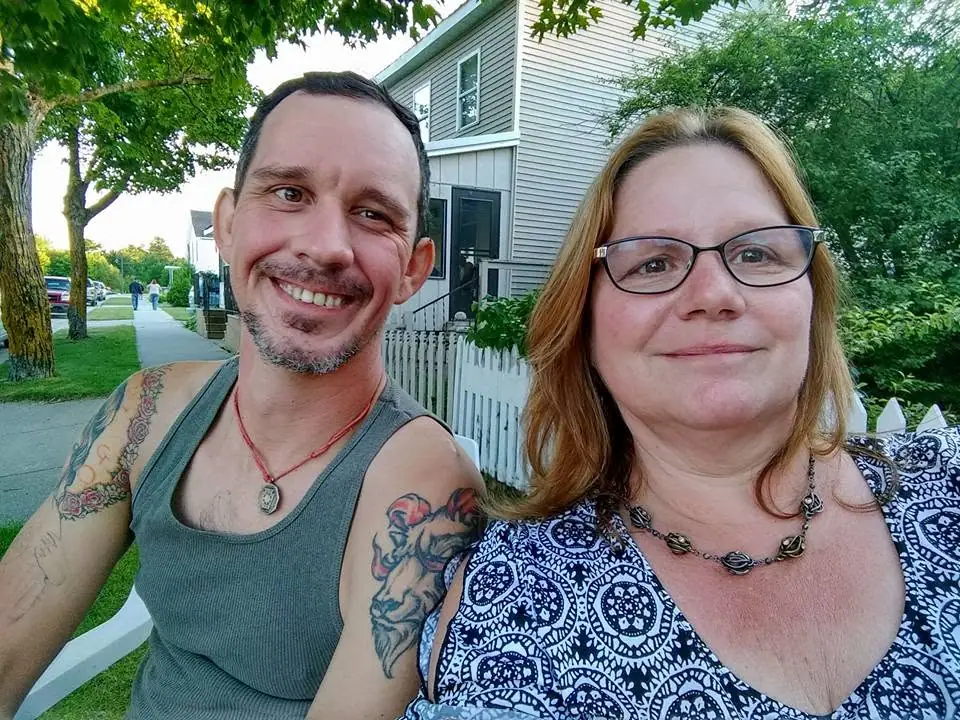 Two smiling adults pose for a selfie beside a house, sidewalk, lawn, and white picket fence.