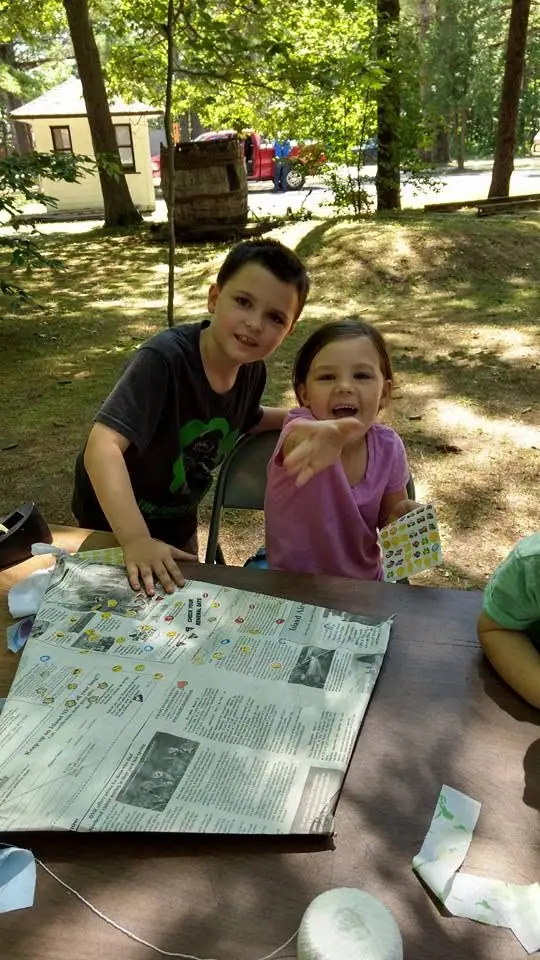 Two children sit at an outdoor table with a large printed paper while another child is partly visible at right.