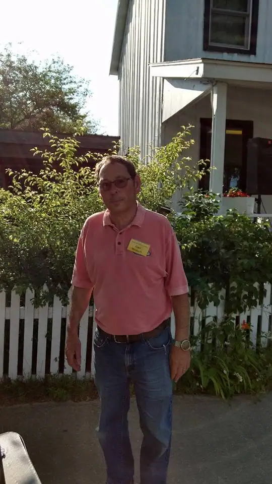 A man wearing a pink polo shirt stands before a house, garden shrubs, and a white picket fence.