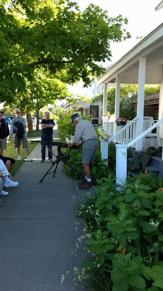 A man adjusts a tripod-mounted firearm-like device beside a porch as several people watch.