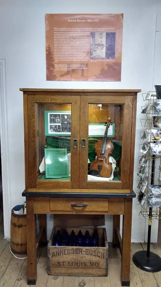 A glass-front wooden display cabinet holds a violin, documents, photographs, and a poster above it.