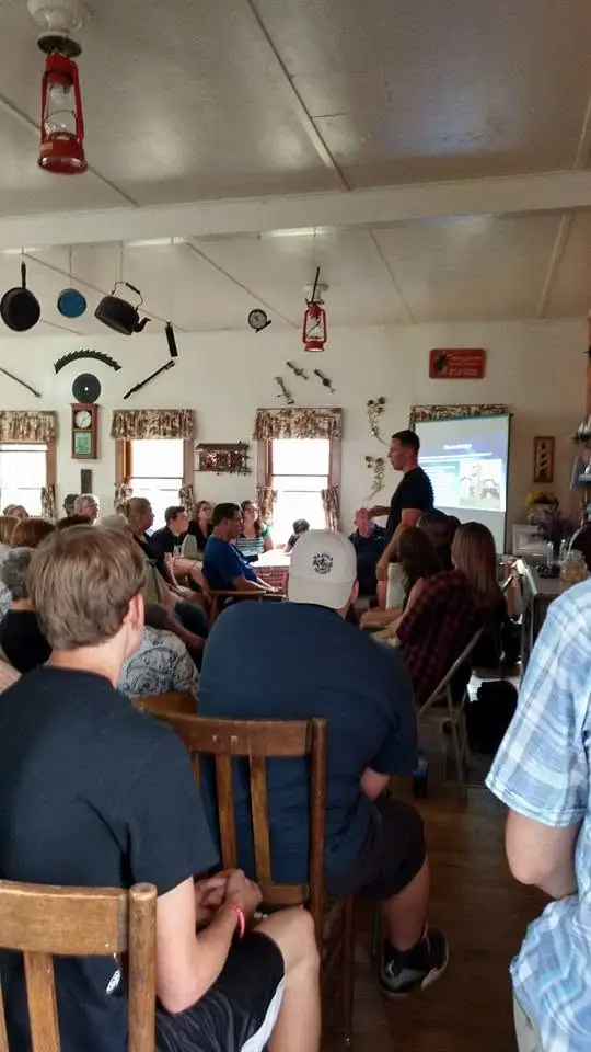 A speaker addresses a seated audience in a room decorated with antique household objects and a projected presentation.
