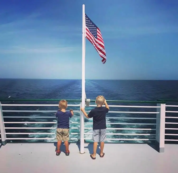 Two children stand at a ship’s stern beside an American flag, looking over the open water and wake.