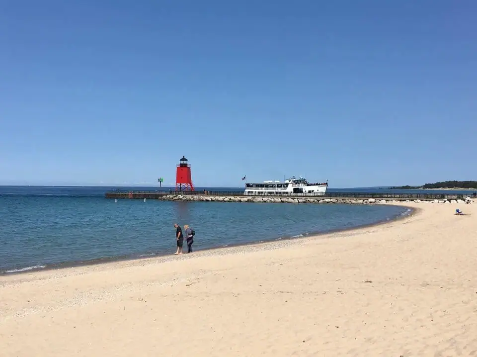 A white passenger ferry passes a red lighthouse on a pier beside a sandy beach.