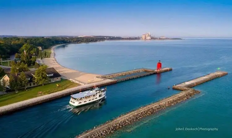 Aerial photograph of a passenger ferry entering a harbor beside a red lighthouse and stone breakwaters.
