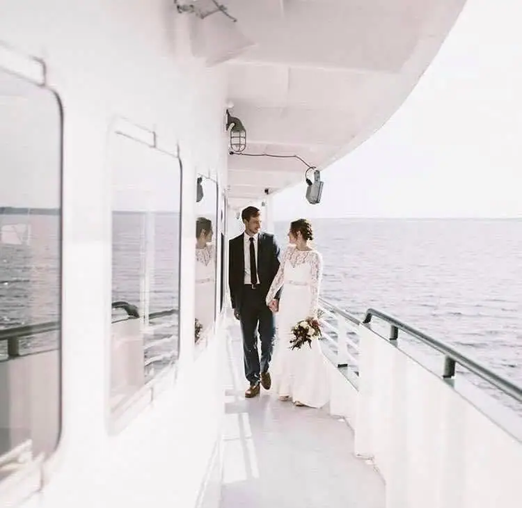 A couple in wedding attire walks along the covered deck of a passenger vessel beside open water.
