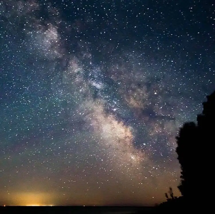 A dense band of the Milky Way stretches across a star-filled night sky above dark shoreline silhouettes.