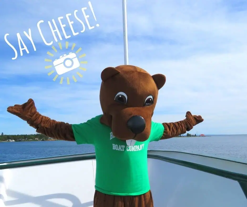 A bear mascot spreads its arms on a boat deck beneath a camera graphic and the words “SAY CHEESE!”
