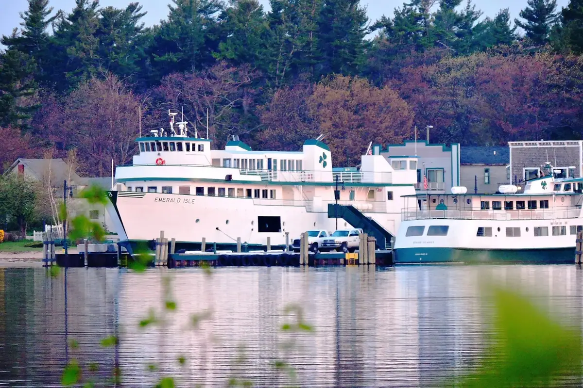 Two white passenger ferries moored at a pier, with pickup trucks, buildings, trees, and calm water.