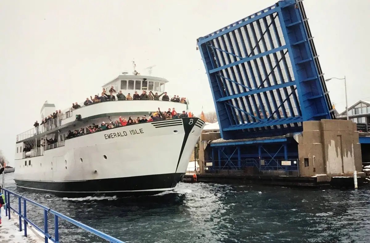 A passenger ferry passes beneath a raised blue drawbridge while people wave from its decks.