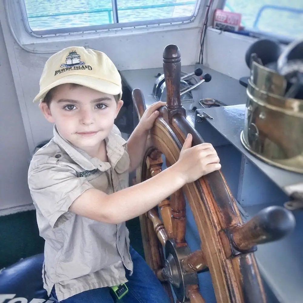 Child wearing a cap reaches for a varnished wooden ship’s wheel inside a boat cabin.