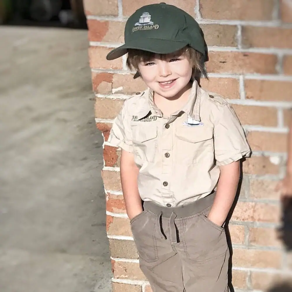 Child in a khaki uniform and green cap stands with hands in pockets beside a brick wall.