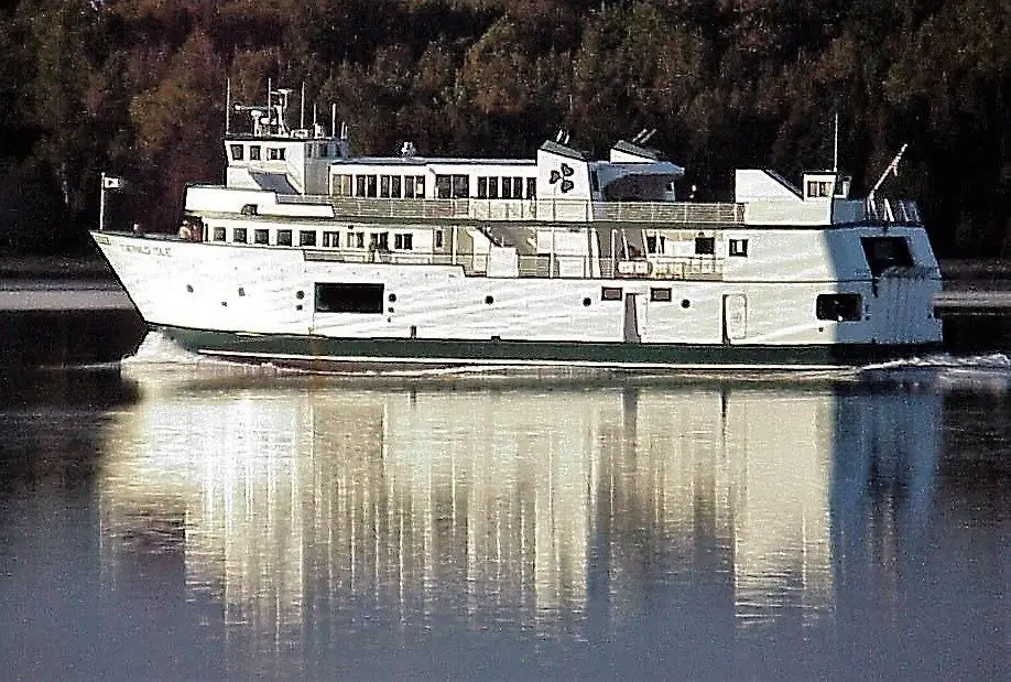 A white multi-deck ferry with a dark green lower hull travels across calm water before a wooded shoreline.