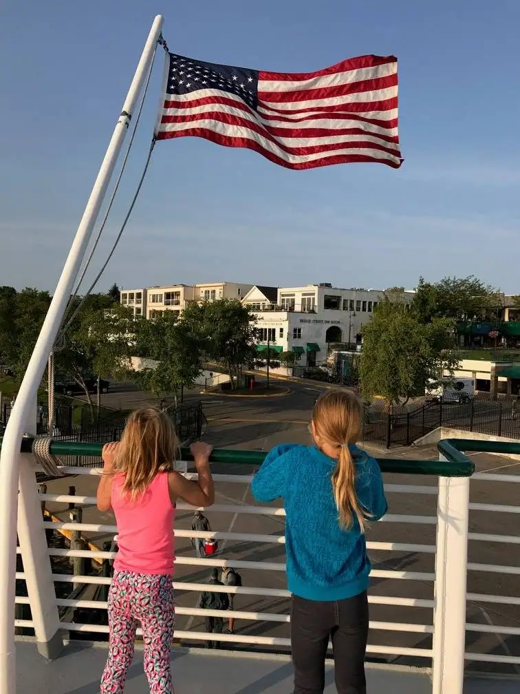 Two children stand at a railing beneath a large American flag, looking toward buildings and streets.