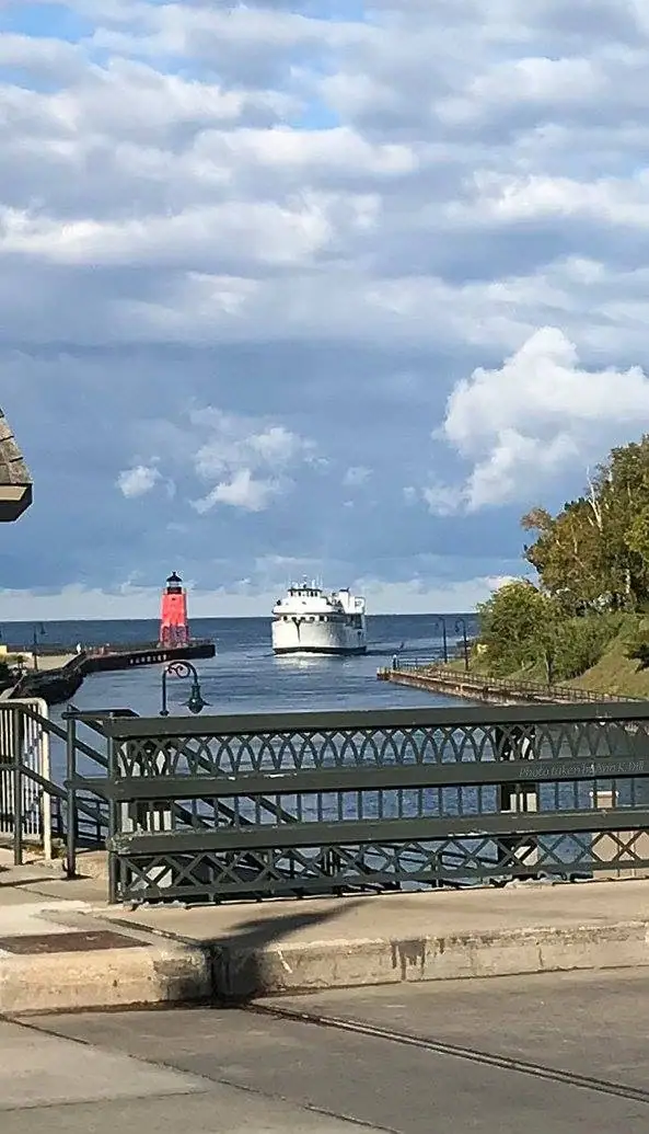 A white ferry approaches a harbor beside a red lighthouse, viewed over a decorative metal railing.