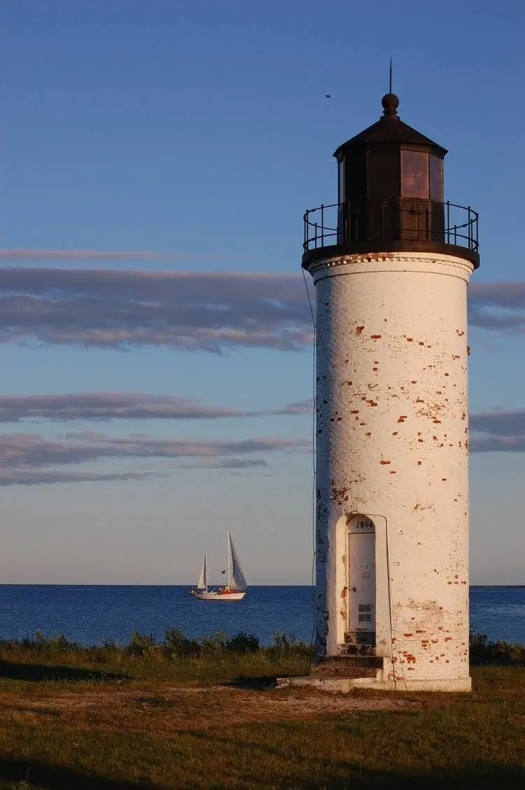 A weathered white lighthouse stands beside the water, with a sailboat visible offshore.