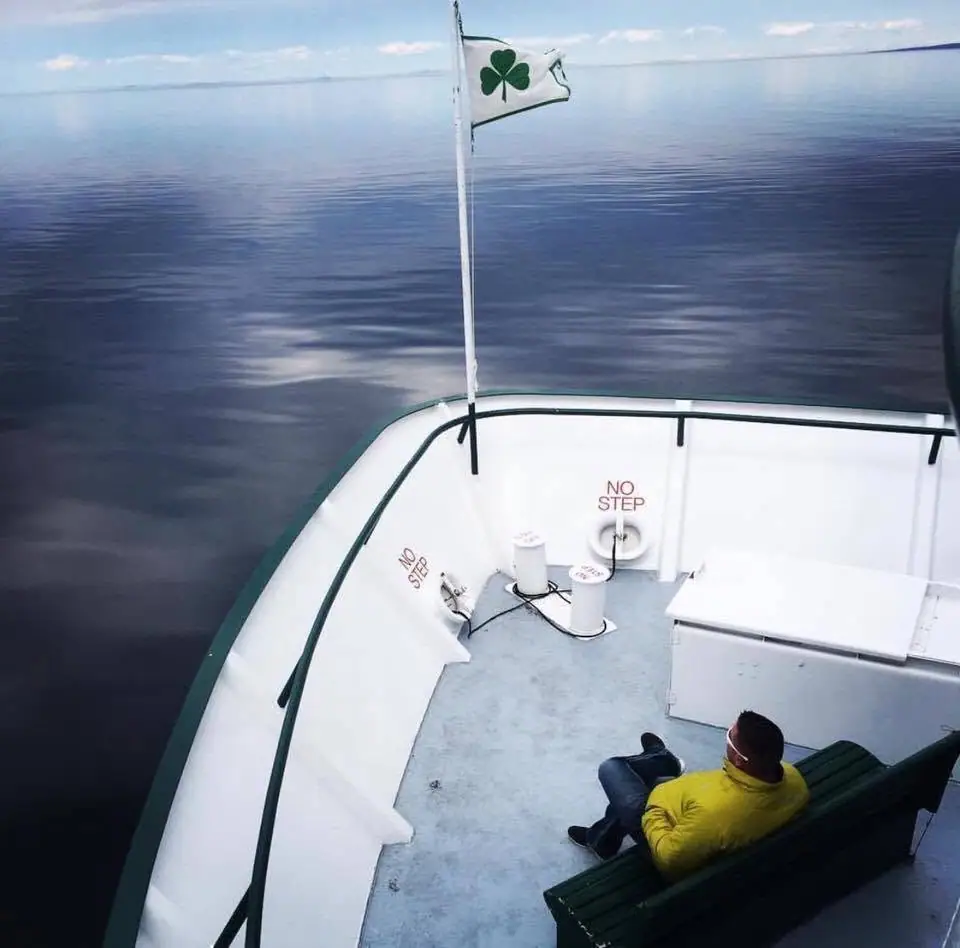 A person in a yellow jacket sits on a green bench on a boat deck beside a shamrock flag.