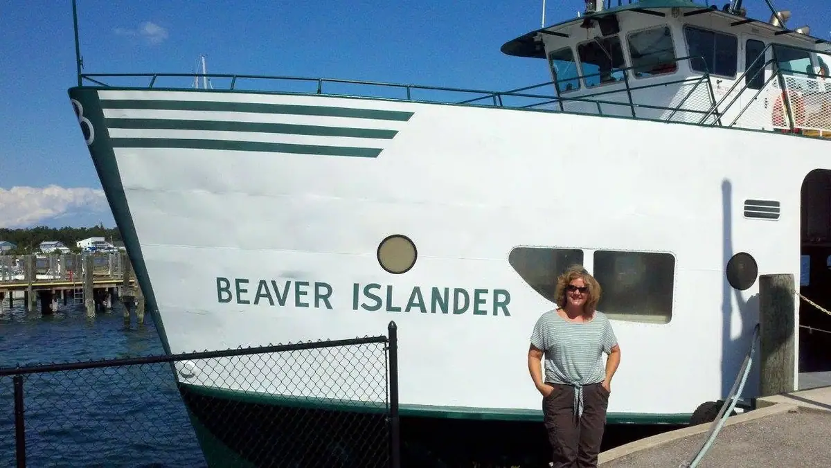 A woman stands beside a white passenger ferry docked behind a chain-link fence.
