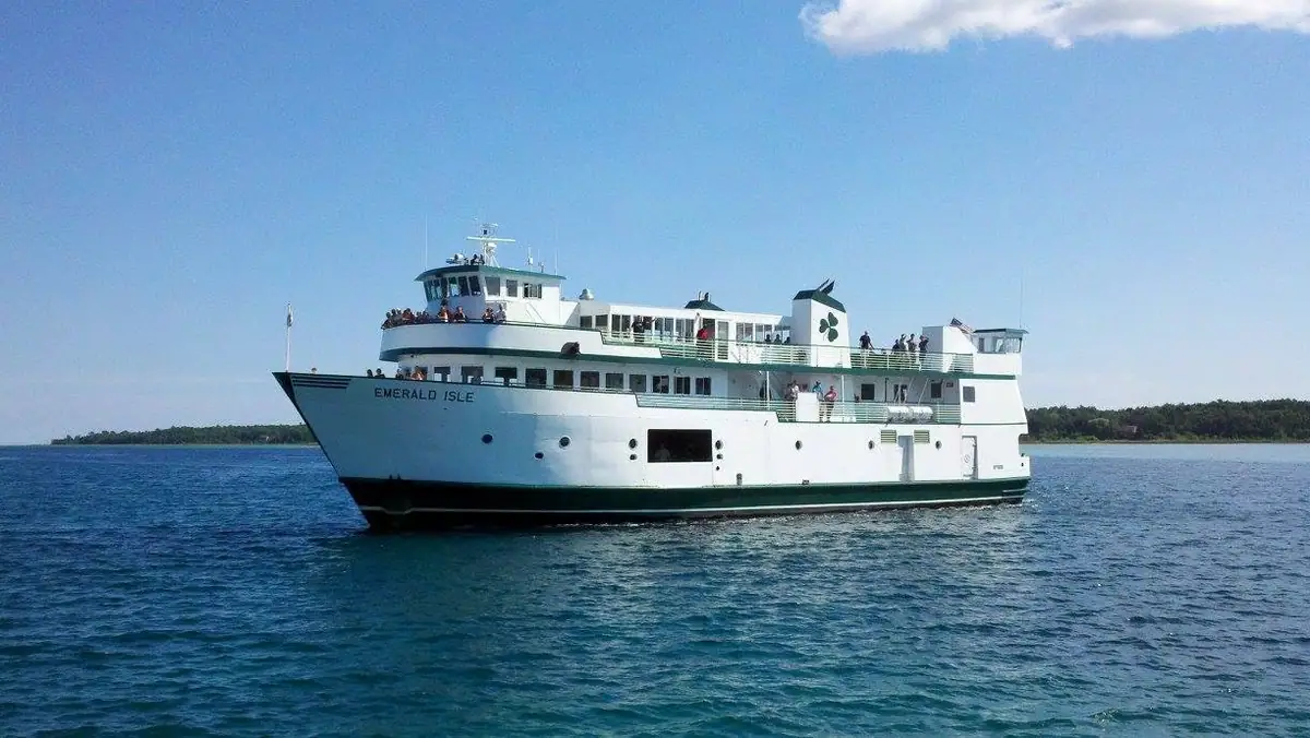 White passenger ferry carrying passengers across blue water, with a wooded shoreline behind it.