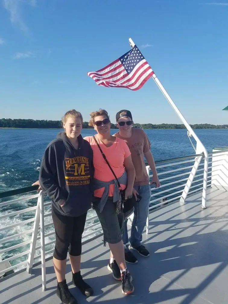 Three people pose on a passenger vessel deck beside an American flag, with open water and a wooded shoreline behind them.