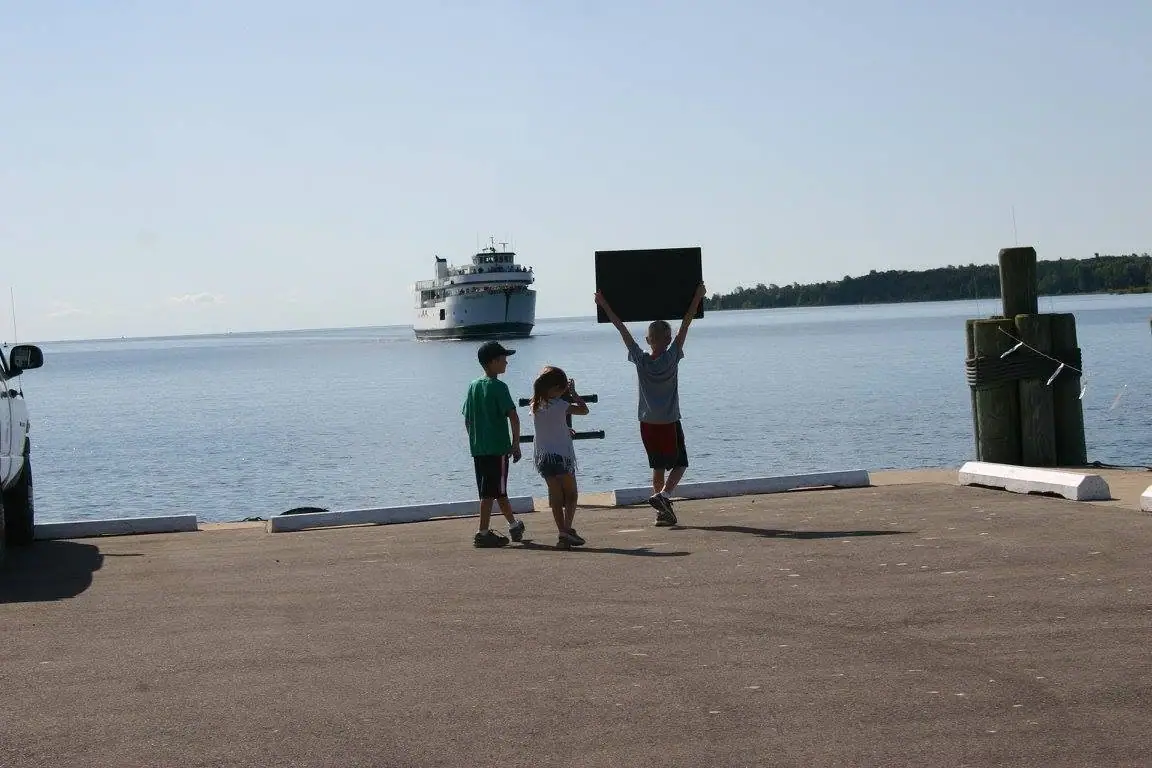 Three children stand on a pier as a ferry approaches across the water.