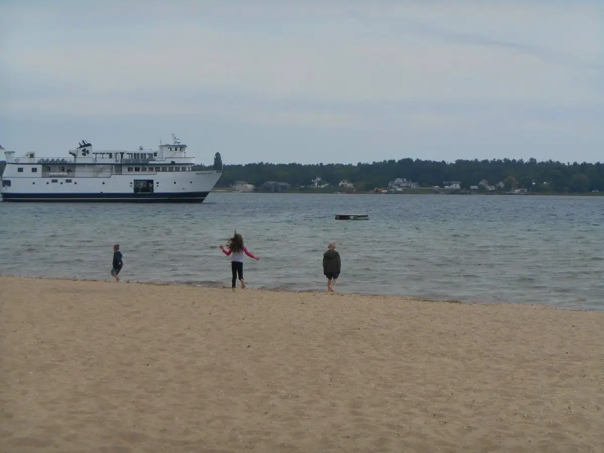 A white ferry passes offshore as three children wade from a sandy beach.