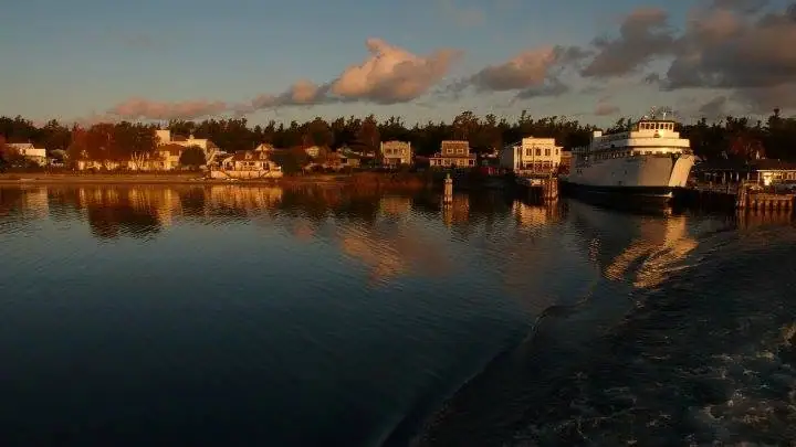 A white passenger ferry is moored beside a shoreline of houses, reflected in calm water beneath warm evening light.