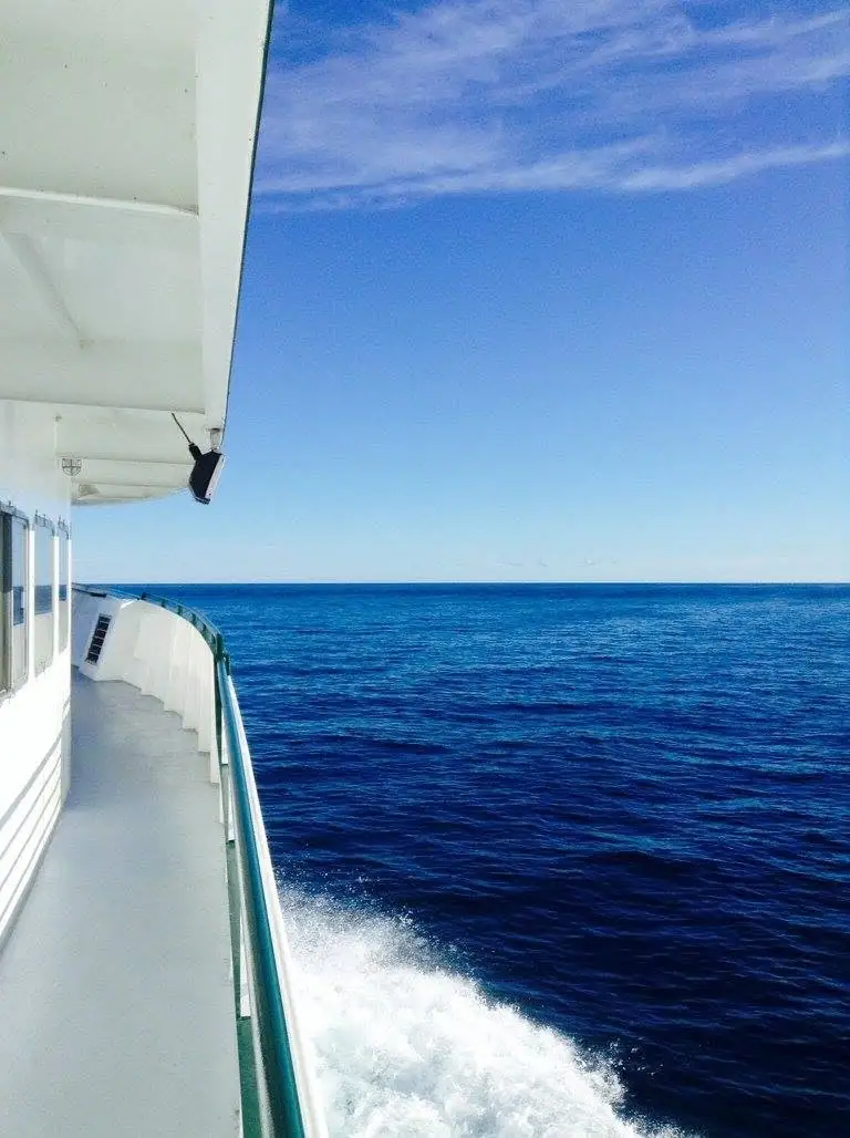 View along a white passenger vessel's side deck, with railing, foaming wake, open blue water, and a partly cloudy sky.