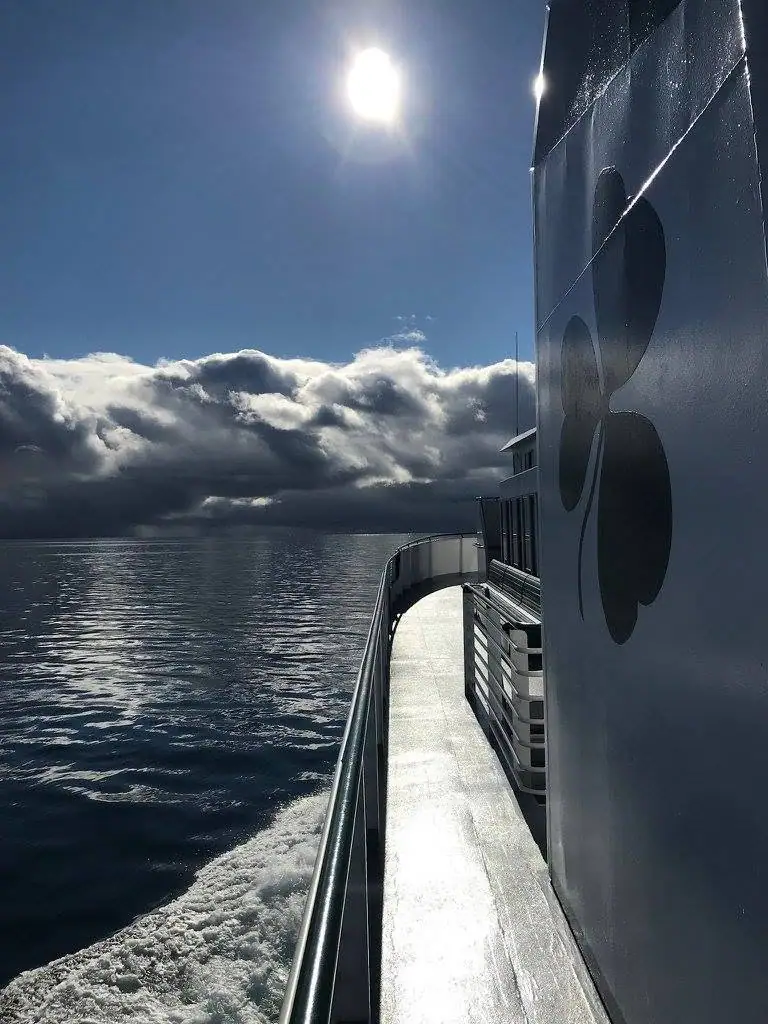 View along a ferry’s side deck toward sunlit water beneath dramatic clouds.