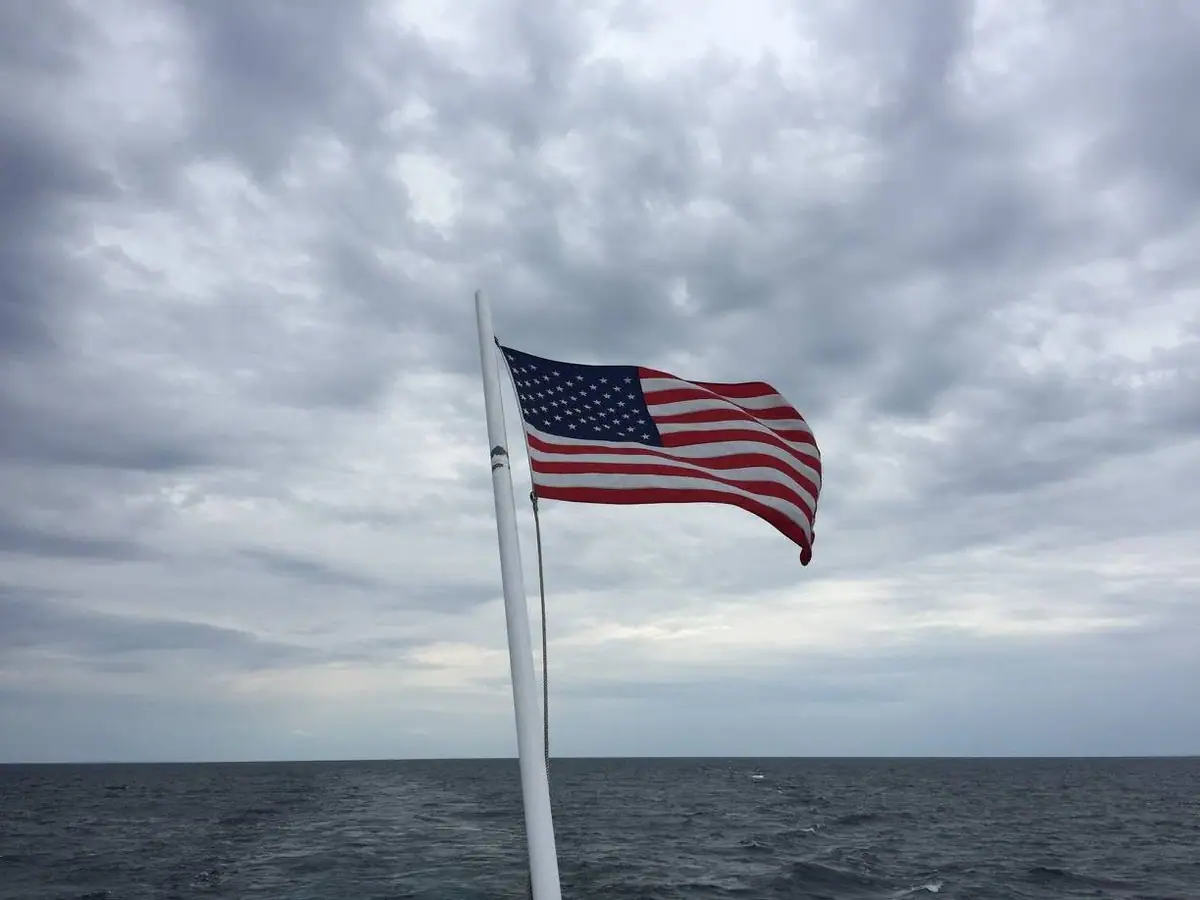 An American flag flies from a white pole above dark, choppy open water beneath a cloudy sky.