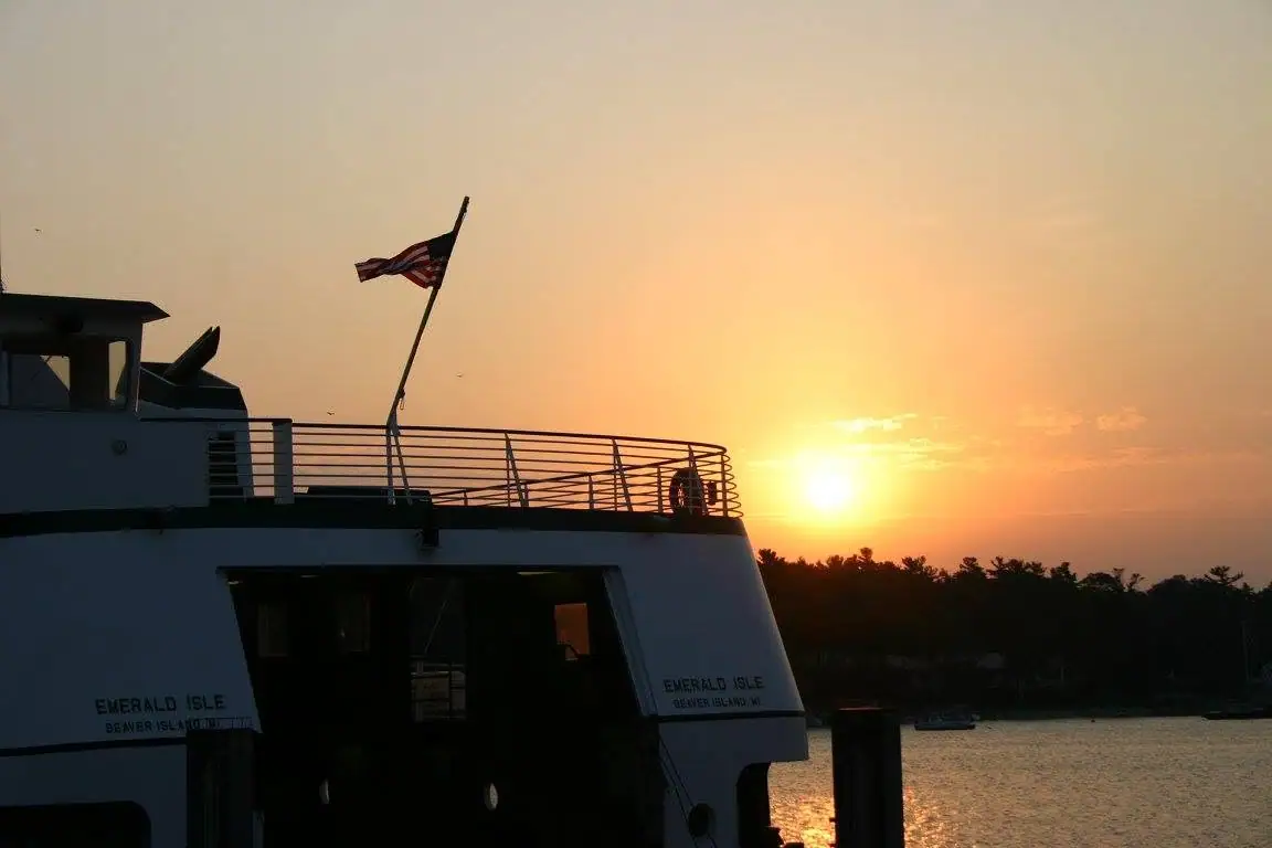 A passenger ferry with an American flag is silhouetted against a bright sunset over the water.