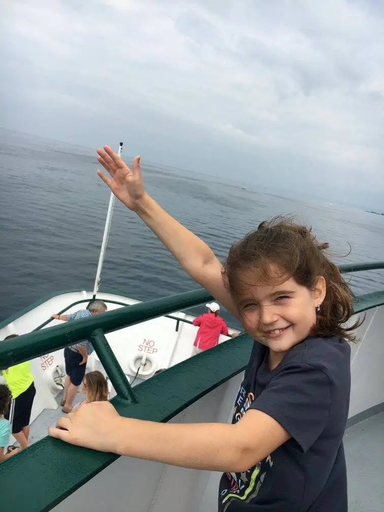 A smiling child waves from a passenger vessel’s deck while other passengers stand near the bow.