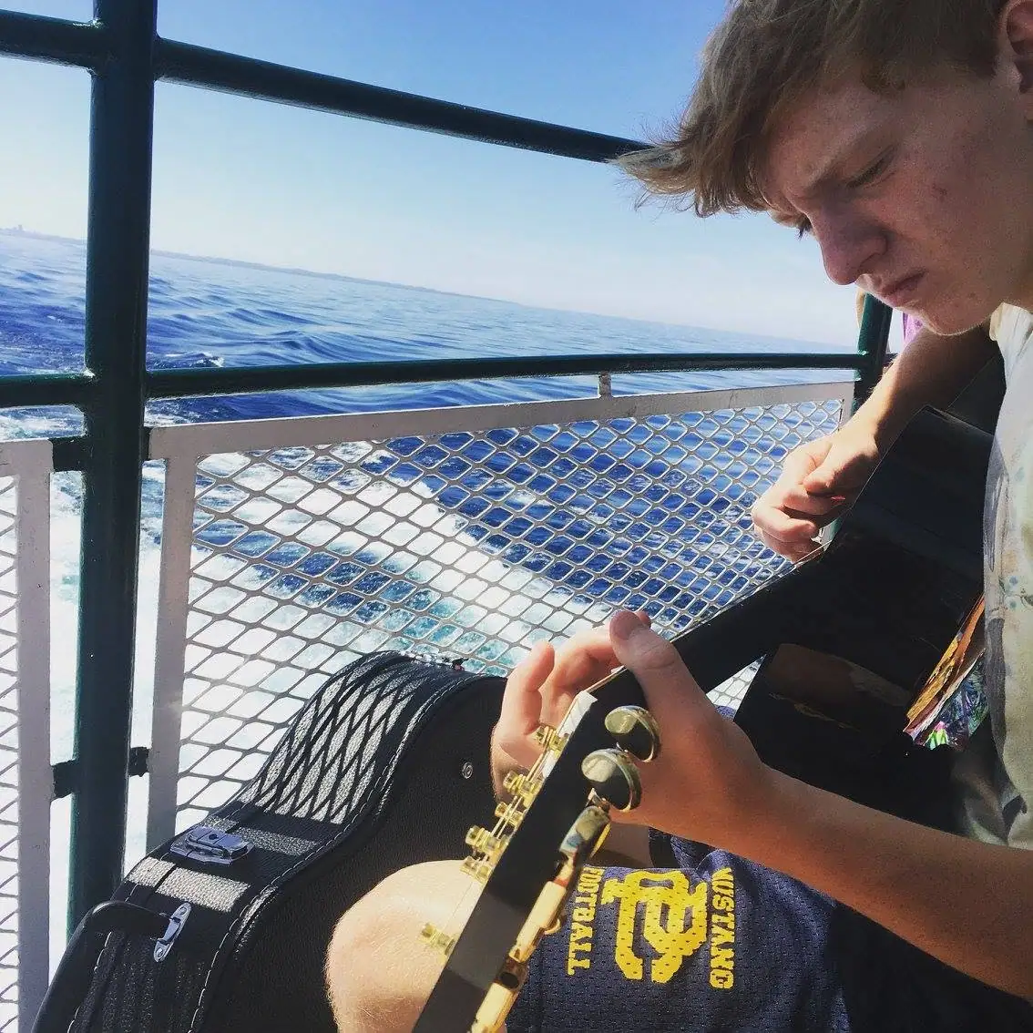 A young person plays a guitar on a boat deck beside the water.