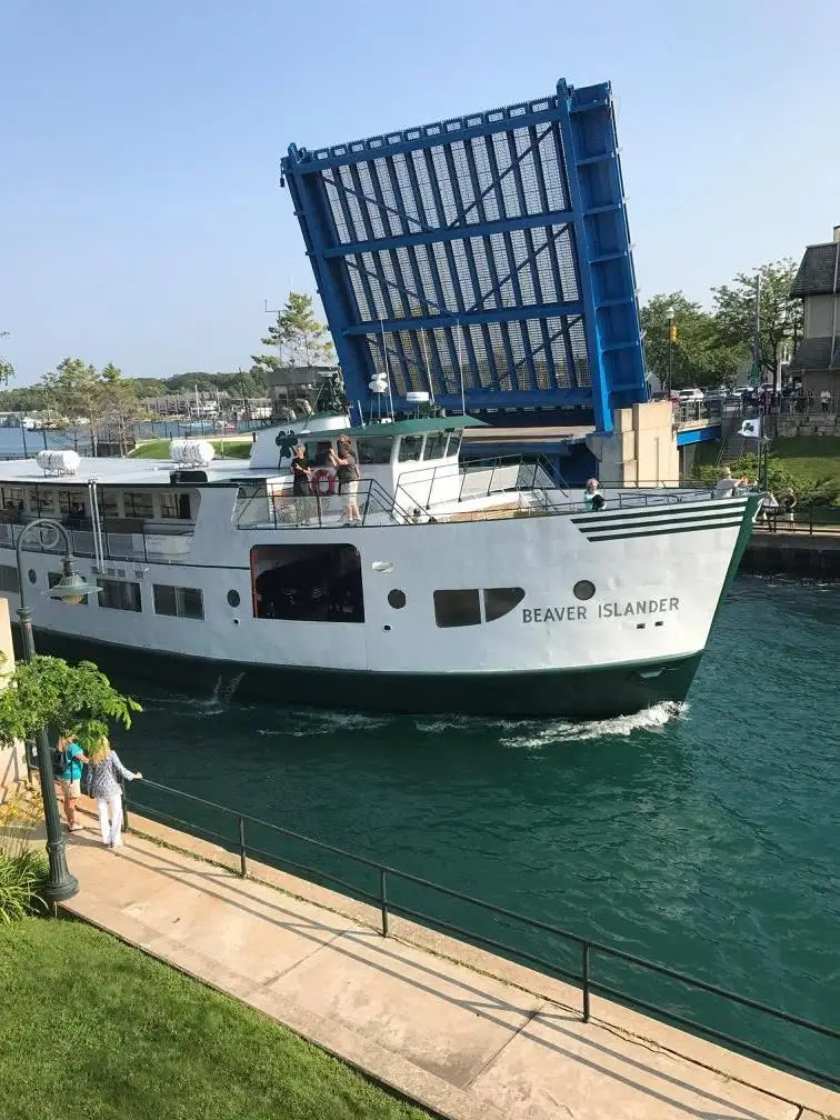 A white ferry passes beneath a raised blue drawbridge while people watch from its deck and the waterfront walkway.