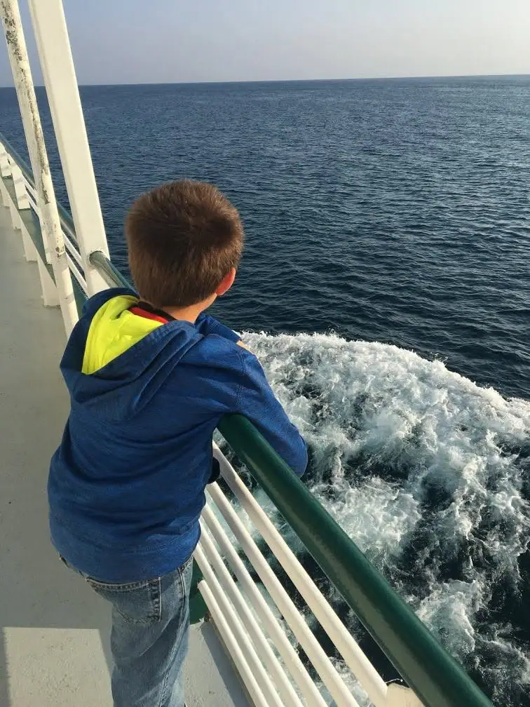 A child leans on a ferry railing above the foaming wake.