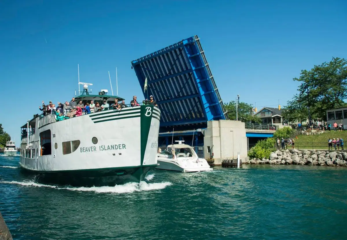 A ferry passes beneath a raised blue drawbridge as passengers gather on deck and people watch from shore.