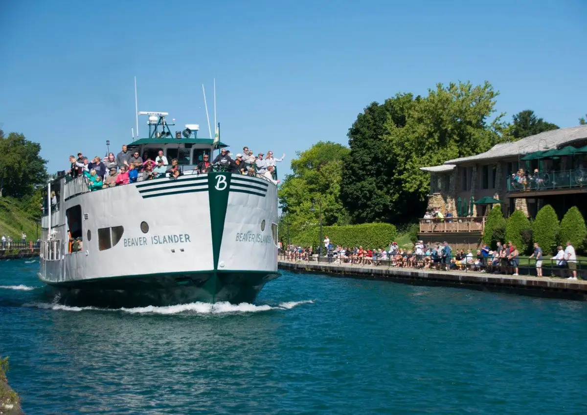 A passenger ferry approaches a crowded pier beside a waterfront building, with people visible on the decks and shore.