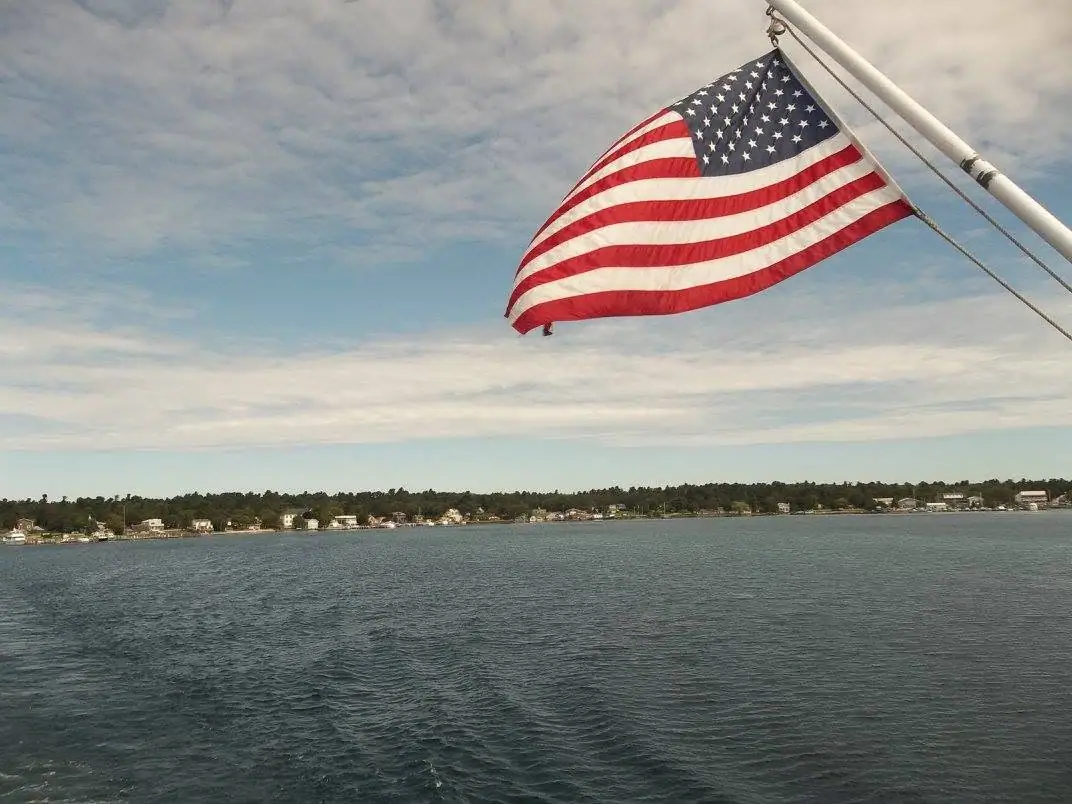 An American flag flies from a boat mast above rippled water and a distant residential shoreline.