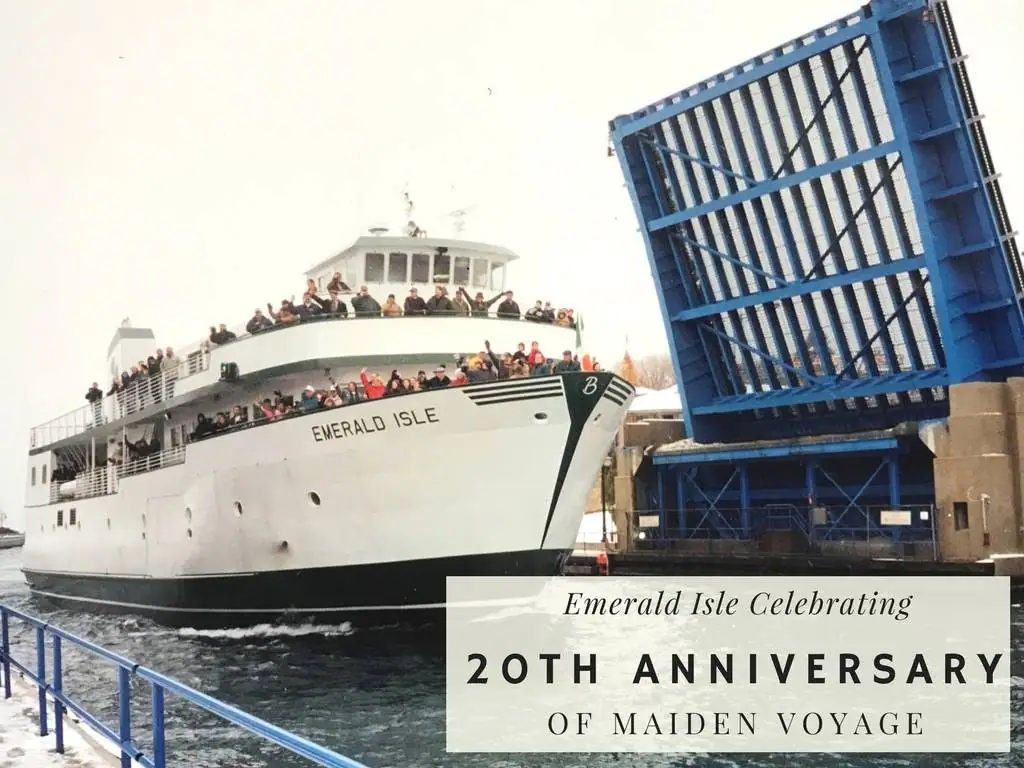 A crowded white ferry passes beneath a raised blue drawbridge beside a waterfront railing.