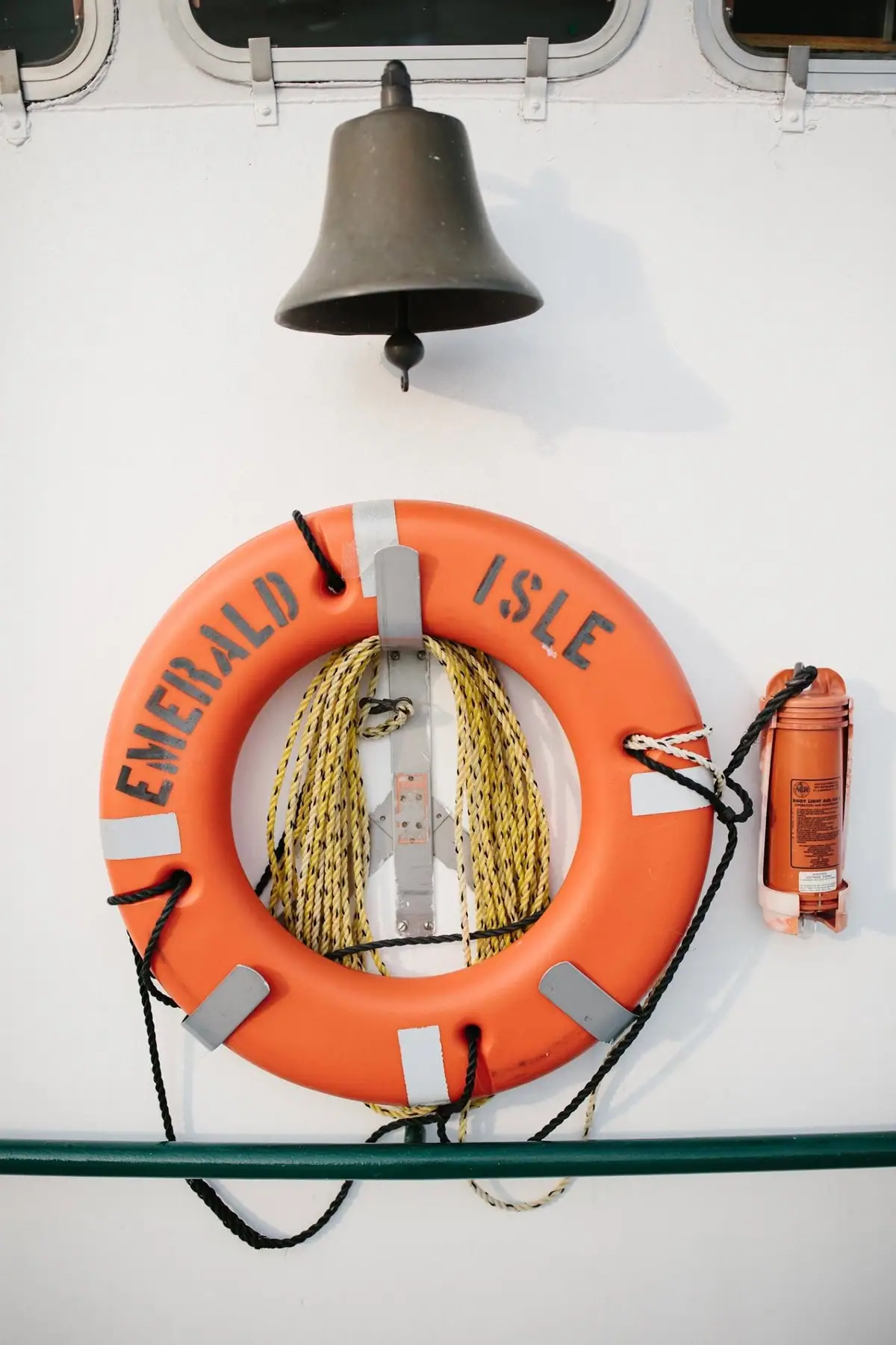 A dark bell hangs above an orange life ring marked EMERALD ISLE on a white wall.