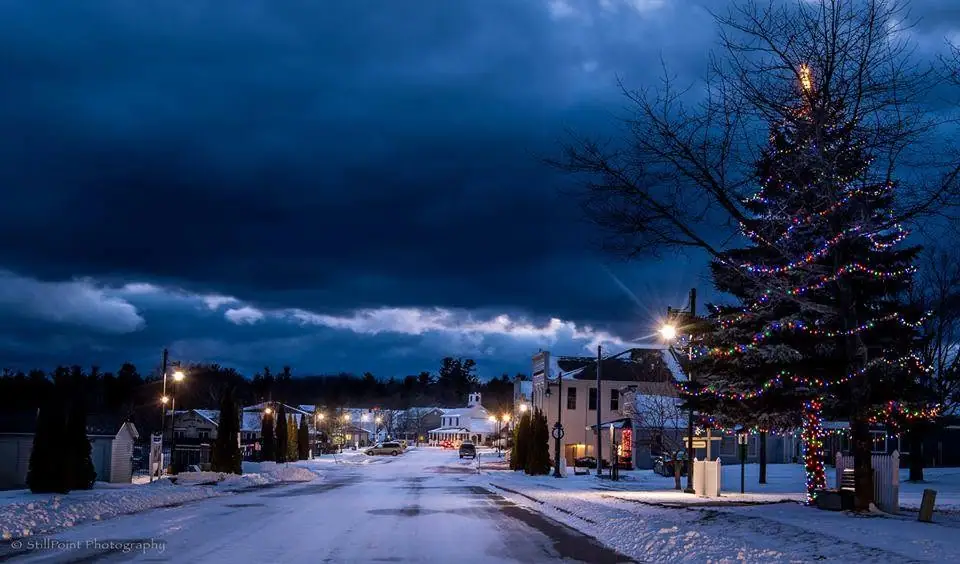 Snow-covered town street at dusk with a brightly decorated evergreen and a church in the distance.