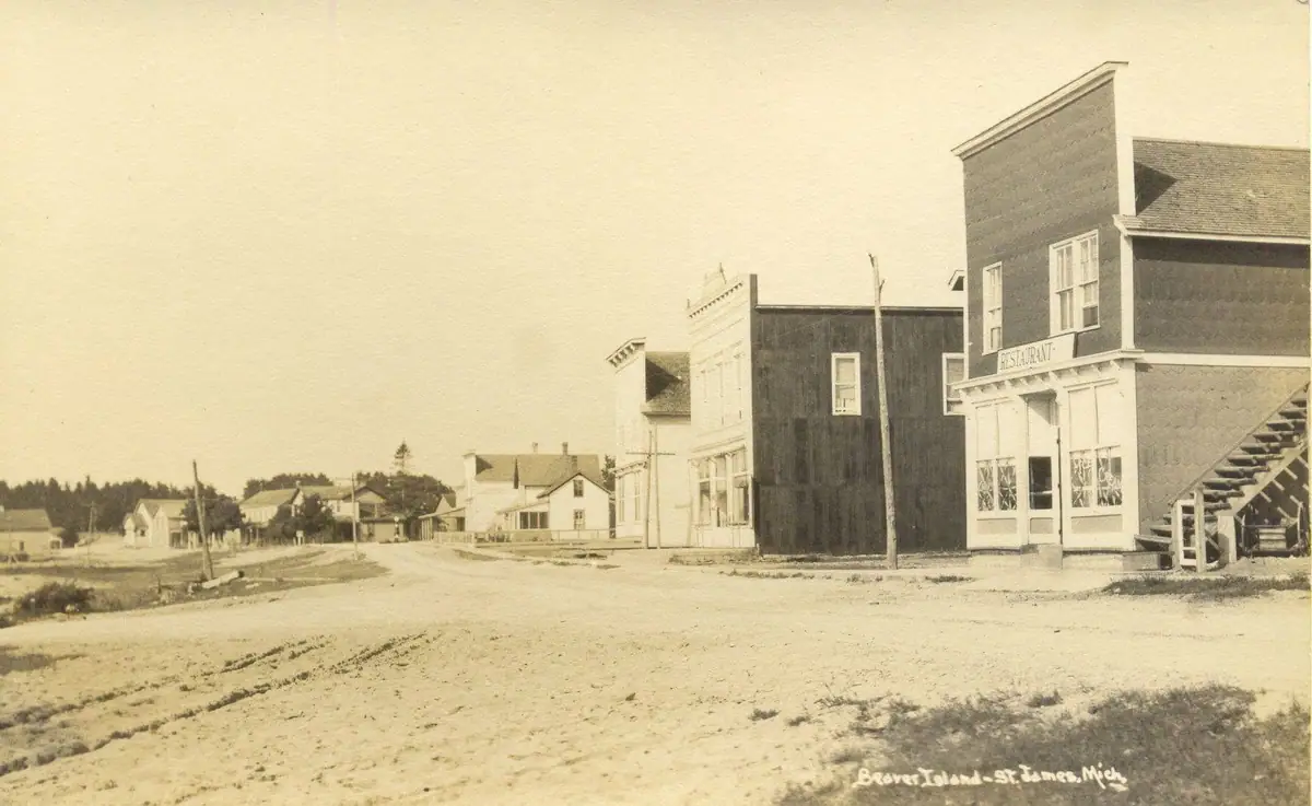 Sepia street scene with an unpaved road, wooden storefronts, homes, and a restaurant sign.