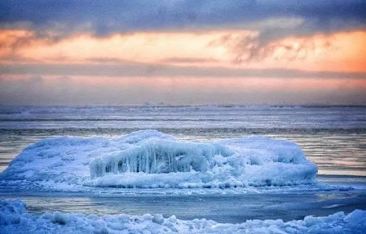 A broad, rounded mass of ice floats on calm water beneath a warm, cloud-filled sky.