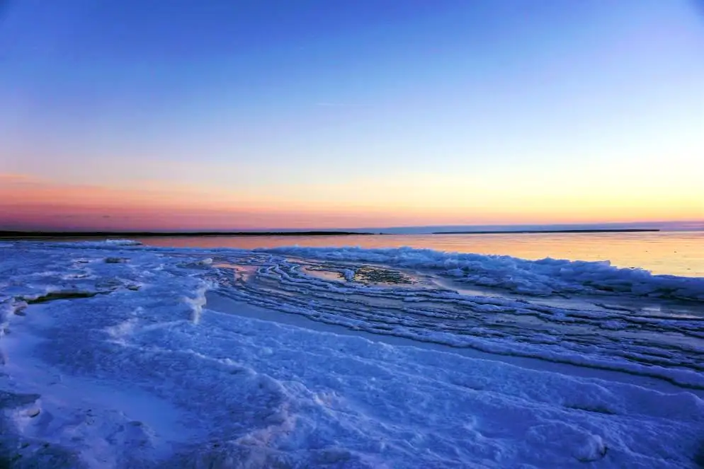 Layered shore ice glows beneath a pastel sunrise, with calm water and a dark horizon beyond.