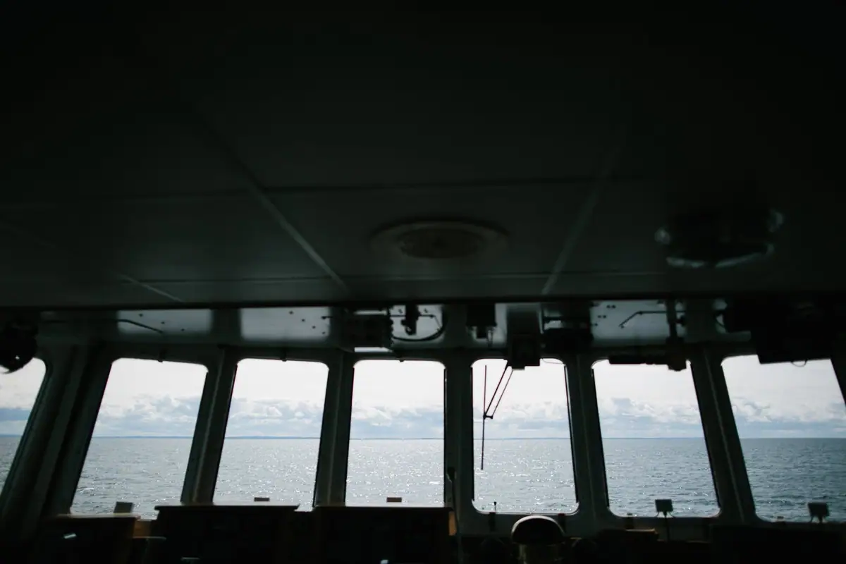View from a vessel's enclosed bridge through five windows toward calm water and a cloudy sky.