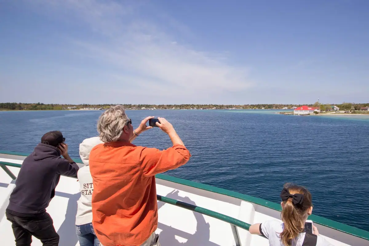 Passengers photograph a wooded shoreline from the open deck of a vessel.