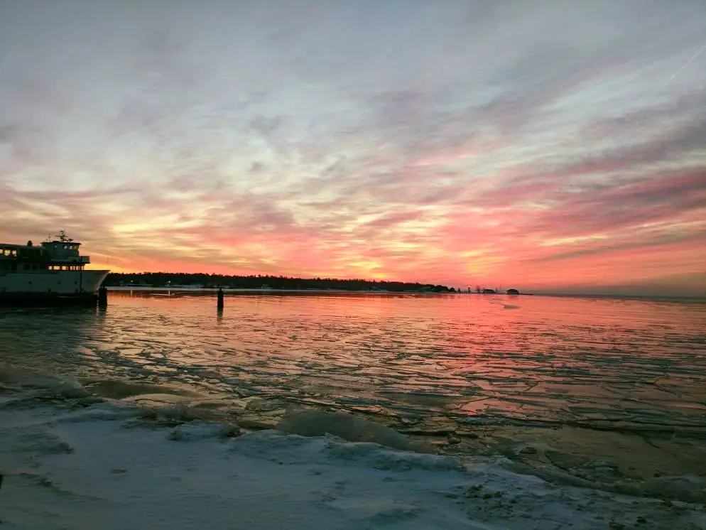 A ferry rests beside a pier as broken ice reflects a vivid pink and orange sunset.