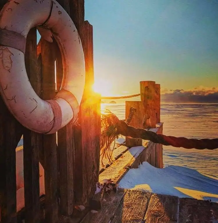 A snow-covered wooden pier with a lifebuoy and rope posts glowing in the low winter sun.