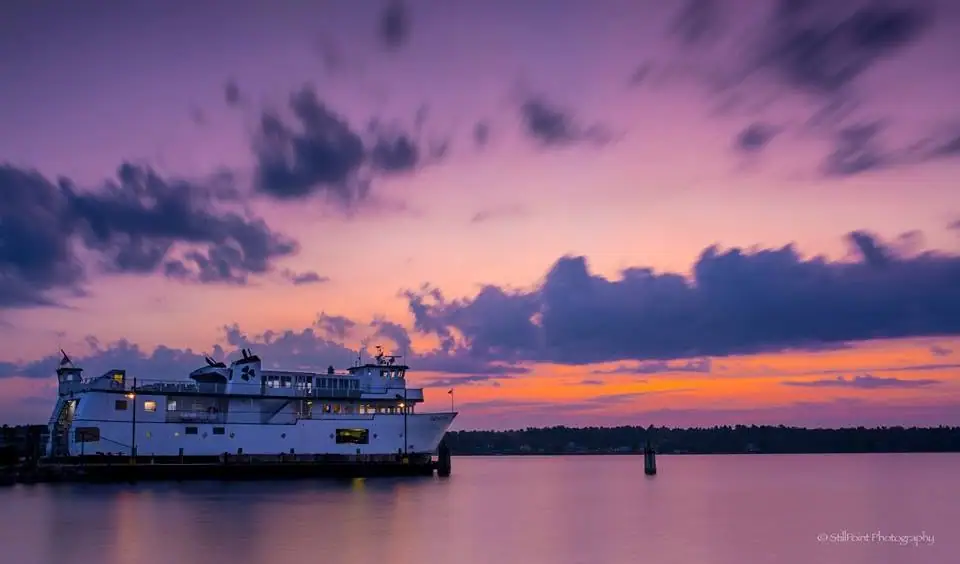 White ferry beside a pier at sunset, with calm water, clouds, and a distant wooded shoreline.
